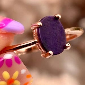 May include: A ring featuring a smooth, oval-shaped deep purple stone set in a polished rose gold-toned metal band. The stone is secured by four small prongs, and the band has a simple, slender design that encircles the finger with a metallic finish.