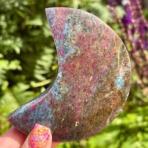 May include: A crescent moon-shaped polished stone carving featuring a mottled texture with vibrant patches of deep magenta, earthy brown, and slate blue mineral inclusions throughout the surface, displaying a smooth and reflective finish.