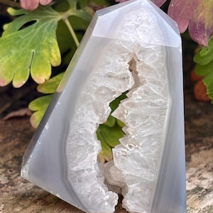 May include: A polished gray agate tower featuring a central vertical geode cavity filled with translucent white crystalline structures. The stone has smooth, banded outer surfaces and a pointed top, showcasing natural mineral formations.