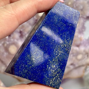 May include: A polished lapis lazuli crystal pyramid in a deep blue hue. The stone features gold flecks, creating a contrast against the blue. The pyramid shape is geometric and contemporary, suitable for display.