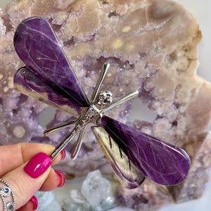 May include: A dragonfly brooch with purple and silver detailing. The wings are made from polished purple stone, and the body and details are silver-toned metal. The brooch is set against a backdrop of a purple crystal.