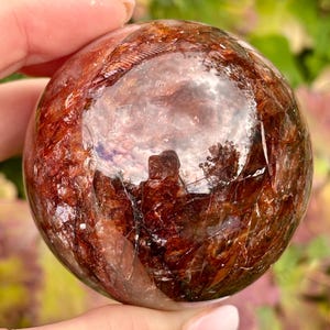 May include: A polished, spherical gemstone featuring deep red and brown inclusions throughout its translucent, crystalline structure. The surface is smooth and reflective, showcasing a complex network of internal fractures and mineral patterns.