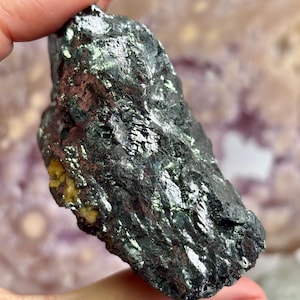 May include: A dark, metallic-looking mineral specimen with a rough, textured surface. The stone has a deep, charcoal gray color with areas that reflect light, creating a shimmering effect. A small section of yellow-brown material is visible.