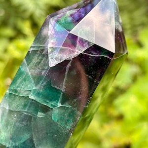 Pode incluir: Uma torre de cristal de fluorita polida com uma ponta hexagonal. A pedra exibe uma mistura translúcida de zonas de cor roxo profundo e verde vibrante com fraturas internas visíveis e inclusões minerais naturais por toda a peça.