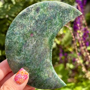 May include: A crescent moon-shaped carving made of green fuchsite stone with visible inclusions of red ruby. The surface features a textured, crystalline appearance with swirling patterns of deep forest green, lighter emerald tones, and small dark flecks.