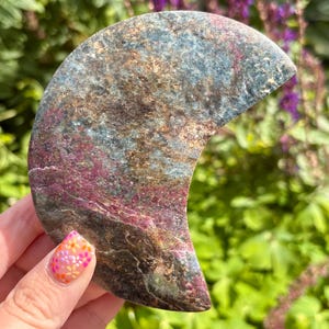 May include: A crescent moon-shaped stone carving featuring a mottled surface with deep magenta, metallic bronze, and slate blue mineral inclusions. The polished rock displays a natural, textured appearance with varied color veins throughout the piece.