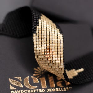 Può includere: Primo piano di un braccialetto di perline nere con un motivo geometrico oro e bianco. Il braccialetto è appoggiato su una scatola nera con la scritta dorata "Scilla HANDCRAFTED JEWELLERY". Il braccialetto è fatto di piccole perline distanziate.