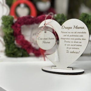 May include: A white wooden heart-shaped sign with a message in Romanian that reads "Draga Mama, Nimeni nu va sti vreodata cat de puternica este dragostea mea pentru tine. Pentru ca doar eu ti-am auzit inima de pe interior. Te iubesc!" The sign is on a white wooden stand.