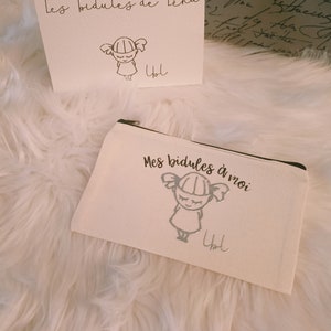 Peut inclure: Deux pochettes en tissu blanc avec un motif argenté d'une fille avec des couettes. Le texte sur les pochettes est "Les bidules de Lena" et "Mes bidules à moi". Les deux pochettes sont signées "Yol".