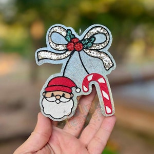 May include: A Christmas ornament featuring a Santa Claus face, a candy cane, and a bow with holly berries. The ornament has a glittery silver finish and is in the shape of a bow with a candy cane and Santa Claus face hanging below.