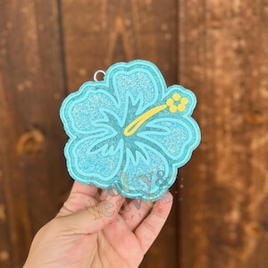 May include: A light blue, glittery hibiscus flower-shaped ornament. The flower has a yellow stamen and a silver loop for hanging. The ornament is held in a hand. The text "calys & leaves craft co" is visible.