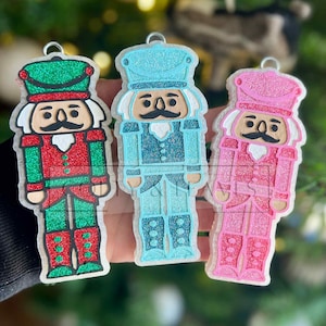 May include: Three glittery Christmas ornaments shaped like nutcrackers. The ornaments are in red, blue, and pink, each with a matching hat and uniform. The nutcrackers have black mustaches and are designed with glitter accents.
