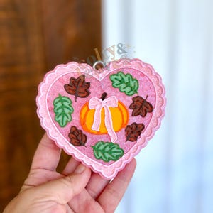 May include: A pink heart-shaped ornament with a scalloped edge. The ornament features a pumpkin with a white bow, surrounded by green and brown leaves. A small silver loop is at the top for hanging.