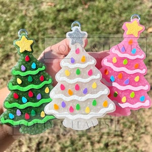 May include: Three Christmas tree ornaments in green, white, and pink. Each tree has colorful lights and a star on top. The green tree has a yellow star, the white tree has a silver star, and the pink tree has a yellow star. Each ornament has a silver loop for hanging.