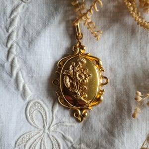 May include: A gold-toned oval pendant featuring a raised relief of a floral bouquet in the centre. The pendant has an ornate, decorative frame with scrollwork details and a small bail at the top for attachment to a chain or necklace.