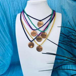 May include: A display of six necklaces with round, copper-colored pendants, each engraved with "02554." The necklaces feature various cord colors, including black, gold, green, and pink, displayed on a white mannequin against a blue backdrop.