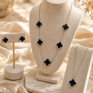 May include: A jewelry set featuring a long silver chain necklace with five black clover-shaped charms, a matching single-pendant necklace, and a pair of stud earrings with black clover-shaped motifs, all displayed on beige velvet stands.