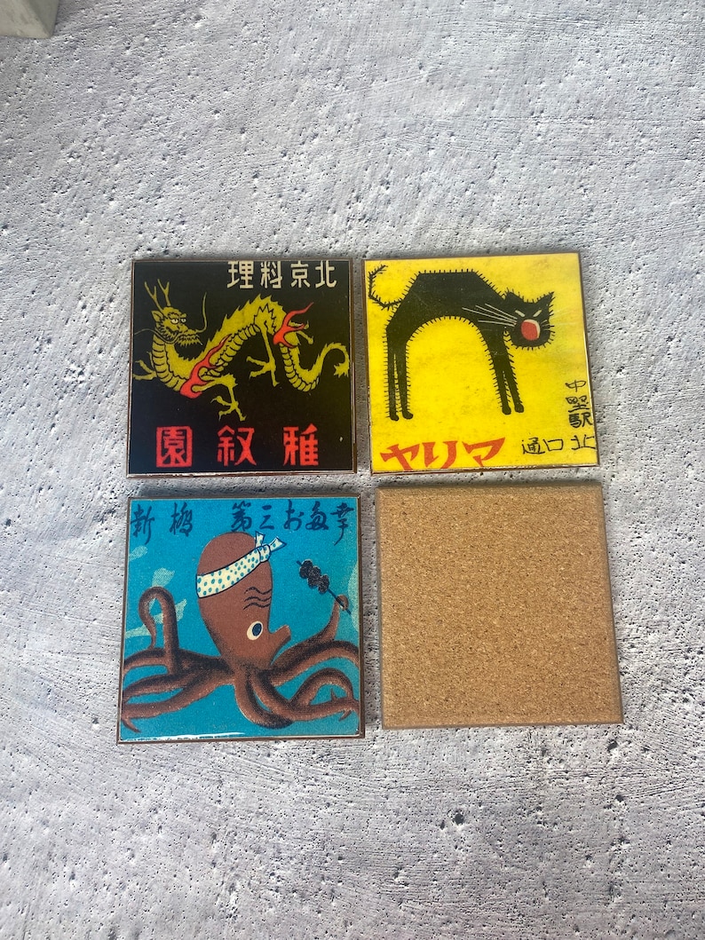May include: Four square coasters with Japanese-inspired designs. One features a black cat on yellow, another a dragon on black, and a third an octopus on blue. The fourth coaster is plain cork.