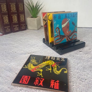 May include: A set of Japanese-themed coasters with a black wooden holder. One coaster features a golden dragon on a black background with red text. Other coasters have colorful illustrations of an octopus and other Japanese imagery.