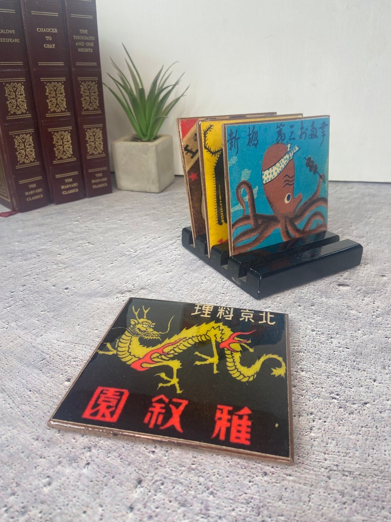 May include: A set of Japanese-themed coasters with a black wooden holder. One coaster features a golden dragon on a black background with red text. Other coasters have colorful illustrations of an octopus and other Japanese imagery.