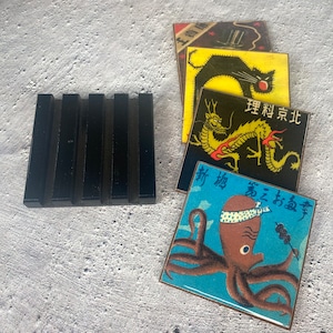 May include: A black soap dish next to a stack of square coasters with Japanese-inspired designs. The coasters feature images of a black cat, a dragon, and an octopus. The octopus coaster has Japanese text.