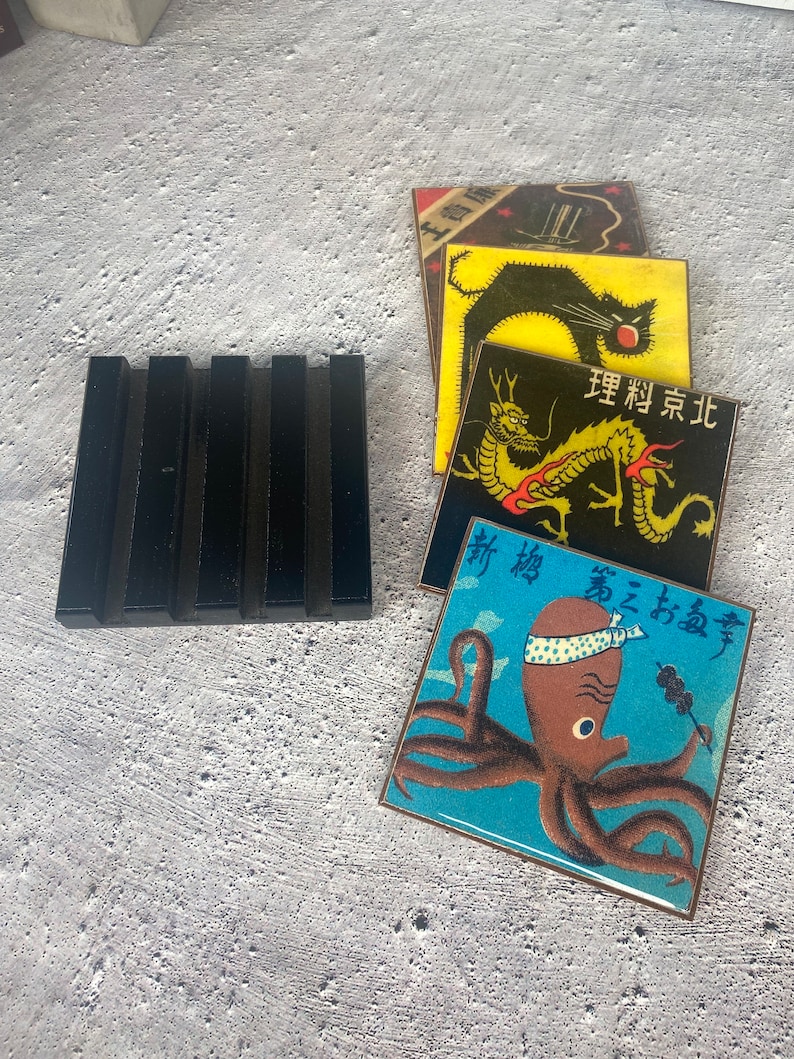 May include: A black soap dish next to a stack of square coasters with Japanese-inspired designs. The coasters feature images of a black cat, a dragon, and an octopus. The octopus coaster has Japanese text.