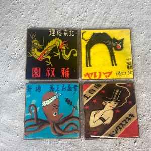 May include: Four square coasters with vintage Japanese-style illustrations. Designs include a yellow dragon on black, a black cat on yellow, an octopus on blue, and a woman in a top hat on brown. Each coaster has Japanese text.
