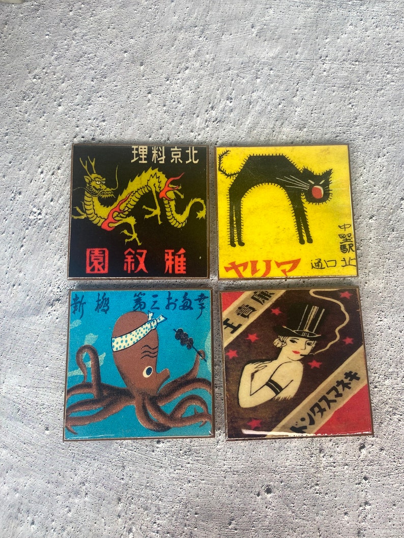 May include: Four square coasters with vintage Japanese-style illustrations. Designs include a yellow dragon on black, a black cat on yellow, an octopus on blue, and a woman in a top hat on brown. Each coaster has Japanese text.