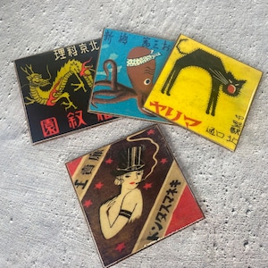 May include: Four square coasters with vintage Japanese-inspired designs. One features a black cat on yellow, another a dragon, an octopus, and a woman in a top hat. Each coaster has Japanese text.