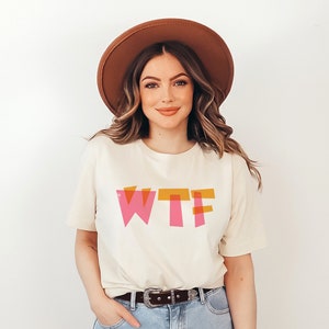 May include: A woman wearing a white t-shirt with the word "WTF" printed in pink and orange letters. She is wearing a brown hat and has long brown hair.