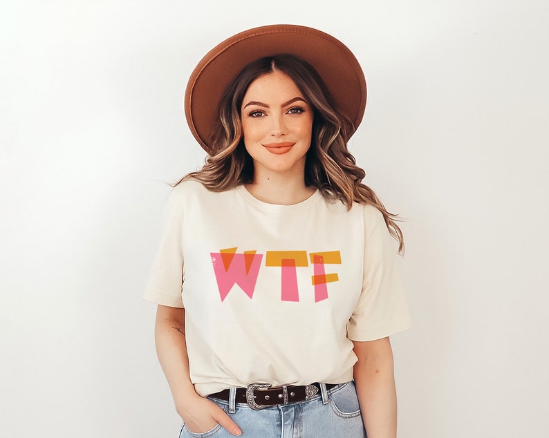 May include: A woman wearing a white t-shirt with the word "WTF" printed in pink and orange letters. She is wearing a brown hat and has long brown hair.
