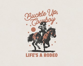 May include: A vintage-style graphic design with a cowboy riding a horse. The text reads "Buckle Up, Cowboy" and "Life's a Rodeo".