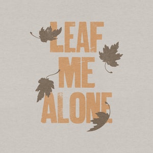 May include: A graphic design with the text "LEAF ME ALONE" in orange letters with a distressed look. There are four brown leaves scattered around the text.