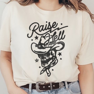 Op de afbeelding: Een wit T-shirt met een zwart grafisch ontwerp van een schedel met een cowboyhoed en de tekst "Raise Hell".
