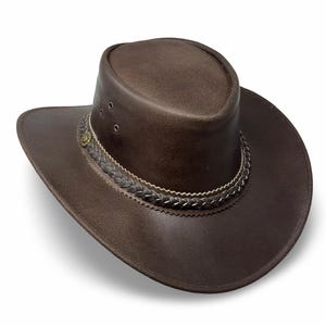 Puede incluir: Un sombrero vaquero de cuero marrón con una banda trenzada y un pequeño botón de metal en la parte delantera.