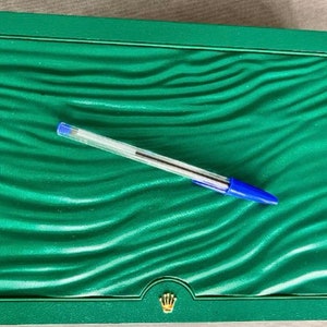 May include: A green, rectangular box with a wave-like pattern on the inside. The box has a gold crown logo on the bottom. A blue pen is lying on the inside of the box.