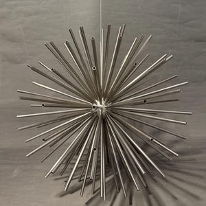 May include: A spherical, metallic sculpture with numerous radiating, hollow tubes. The tubes vary in length and are arranged to create a spiky, starburst-like appearance. The sculpture is suspended by a thin wire against a gray backdrop.