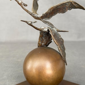 Puede incluir: Escultura de bronce que representa dos pájaros en vuelo, posados sobre una esfera de bronce. Las aves tienen las alas extendidas y están situadas encima de la esfera. La escultura está montada sobre una base cuadrada.