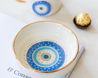 May include: A white ceramic bowl with a blue and gold evil eye design. The bowl has a gold rim and is sitting on a magazine.