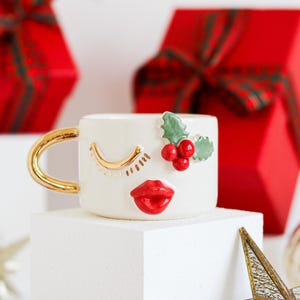 Puede incluir: Taza de cerámica blanca con asa dorada y una cara caprichosa. La cara tiene los ojos cerrados con pestañas doradas, un labio rojo y una rama de acebo verde con bayas rojas.