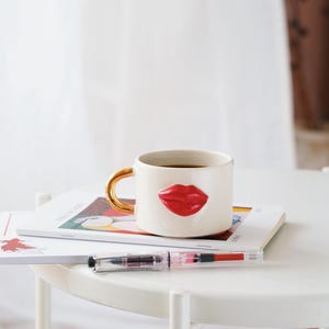 Pode incluir: Caneca de cerâmica branca com alça dourada e design de lábios vermelhos em 3D. A caneca está em cima de uma pilha de revistas em uma mesa branca.