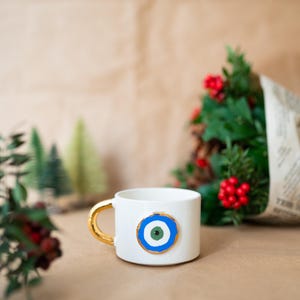 May include: White ceramic coffee mug with a gold handle and a blue and green evil eye design.