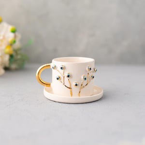 Puede incluir: Una taza de té y un platillo de cerámica blanca con un diseño de árbol dorado y detalles azules de ojo turco.