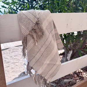 Puede incluir: Una manta tejida a rayas beige y blancas con flecos, colgada sobre una valla de madera blanca. La manta tiene un aspecto texturizado y probablemente está hecha de fibras naturales. El fondo presenta follaje verde y una playa arenosa.