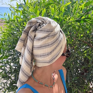 Puede incluir: Una toalla a rayas beige y negras envuelta alrededor de la cabeza. La persona lleva un traje de baño azul y un collar de cuentas turquesas. Se usan gafas de sol. El follaje verde y el mar están en el fondo.