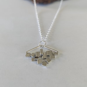 May include: A silver-toned necklace with a geometric pendant. The pendant features a triangular top with a cluster of small, square and rectangular shapes hanging below. The chain is delicate and the pendant is centered.