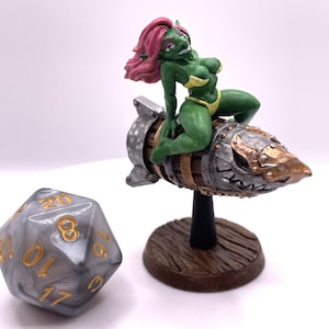 May include: A green and pink figurine of a female goblin riding a rocket-shaped object. The rocket is made of metal and wood and has a detailed design. The goblin is wearing a yellow bikini and has long pink hair.