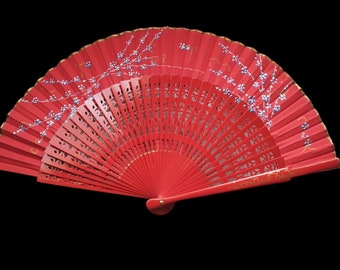 May include: A vibrant red hand fan with a floral design. The fan features a painted branch with white and blue flowers and gold accents. The fan is made of wood and has a decorative, openwork design.