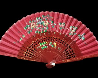 May include: A red hand fan with a floral design. The fan is painted with flowers in shades of yellow, green, and blue. The fan's wooden handle is a darker shade of red and has decorative cutouts.