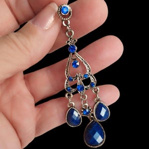 May include: Silver-toned chandelier earring with multiple blue gemstones. The earring features a circular top, a heart-shaped design, and three dangling teardrop-shaped gems. The blue stones are faceted, catching the light.